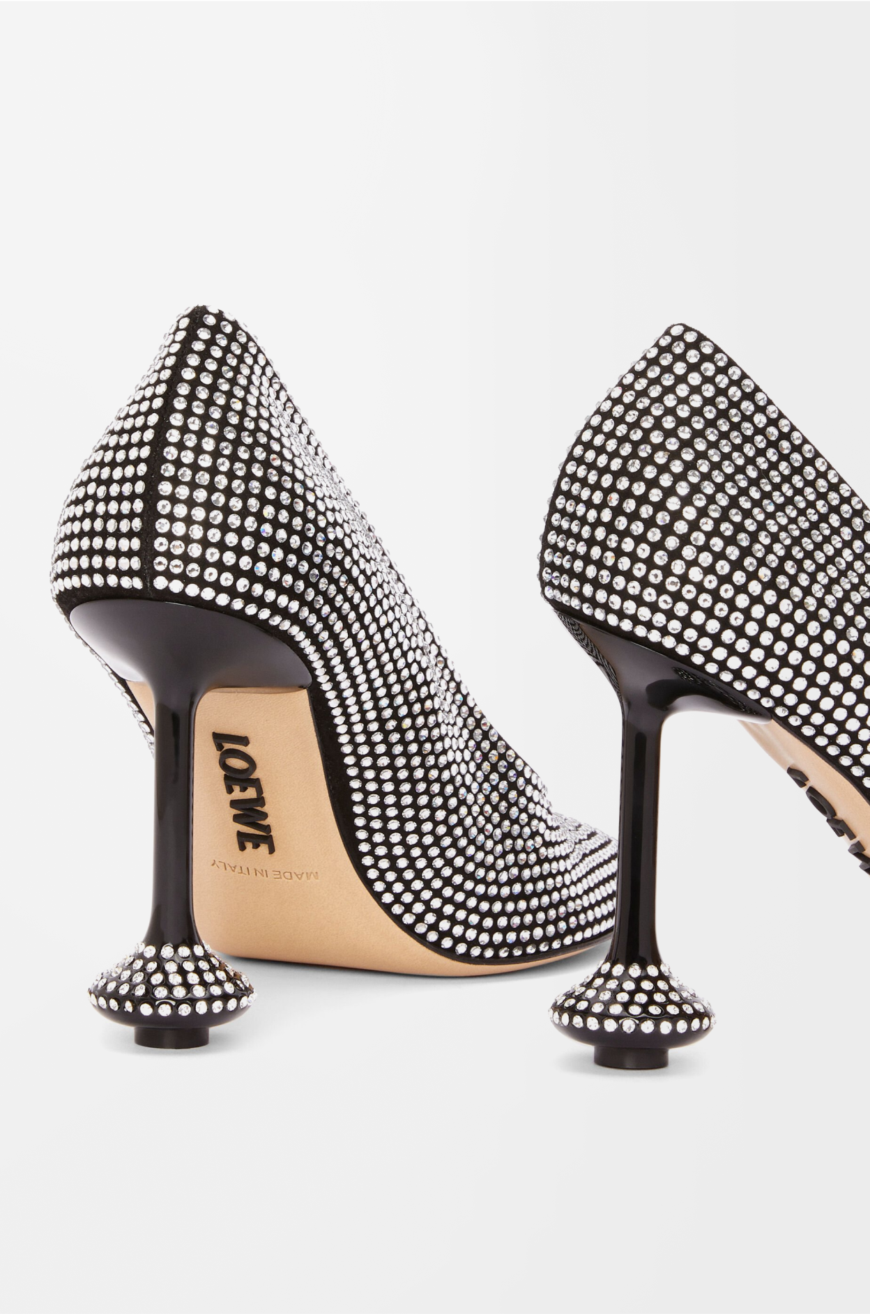 A close-up, angled view of a pair of Loewe "Toy pump" shoes. The black shoes are completely covered in small, sparkling rhinestones. The unique sculptural heel is visible, with the word "LOEWE" subtly placed on the sole.