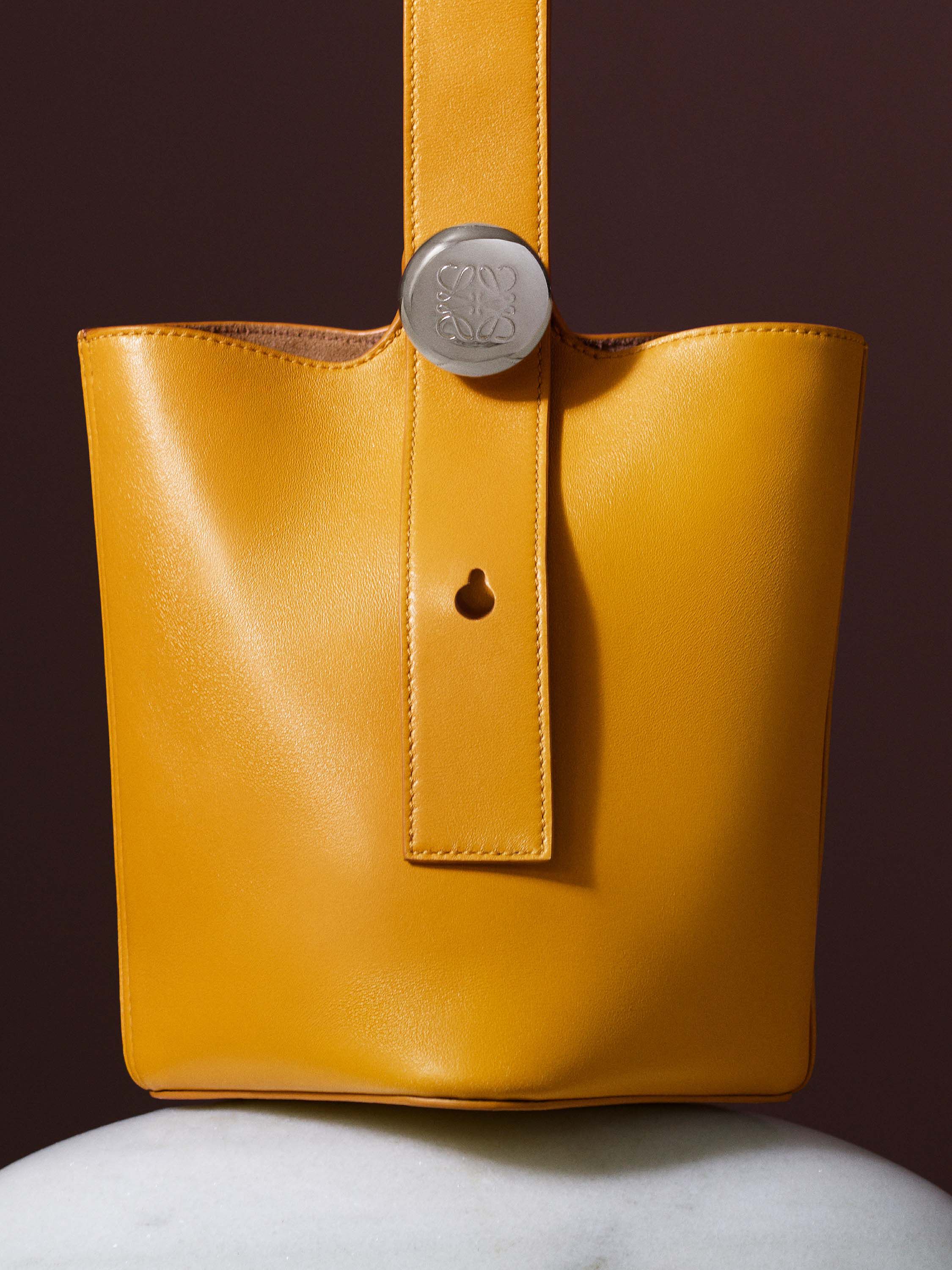 A close-up, professional studio shot of a shiny, yellow leather Loewe bucket bag. The bag has a minimalist strap with a silver "Anagram" button and a small circular cutout.