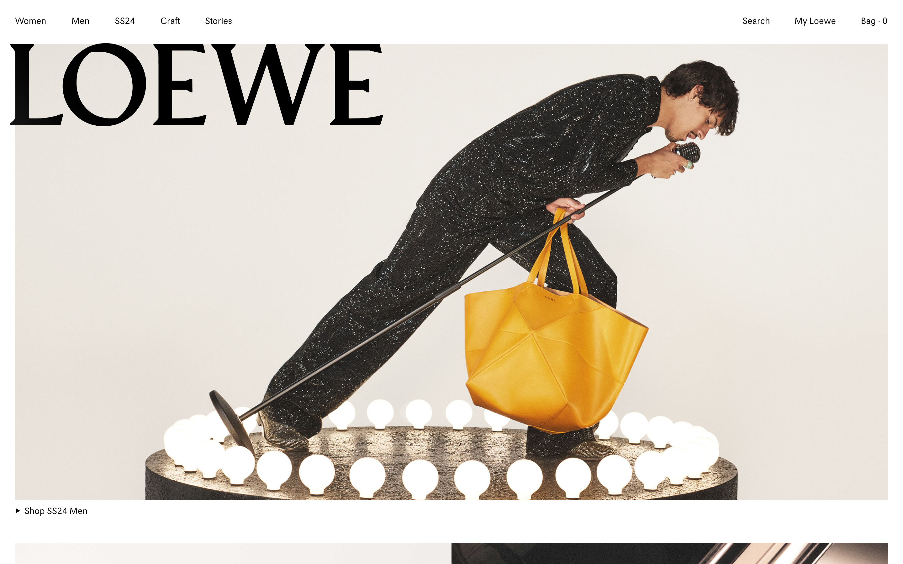 A vibrant landing page for Loewe's SS24 collection. A male model in a shimmering black outfit, holding a microphone, kneels on a lighted stage. He is holding a large, mustard-yellow "Puzzle Fold" tote bag.